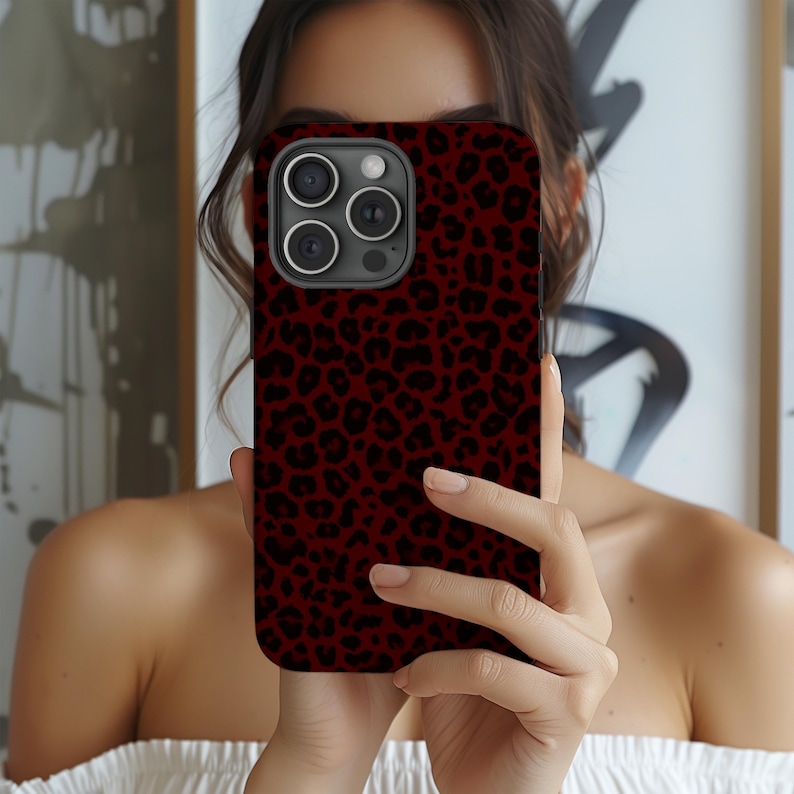 May include: A smartphone case with a dark red background and a black leopard print pattern. The case has a camera module with three lenses and a flash. The phone case is held up in front of a person.