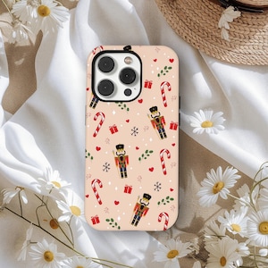 Może przedstawiać: Etui na telefon z motywem świątecznym. Wzór powtarza się z dziadkami do orzechów, cukrowymi laskami, pudełkami na prezenty, płatkami śniegu i sercami na jasnym, brzoskwiniowym tle. Zaprojektowane dla smartfona.