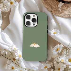 May include: Sage green phone case featuring a minimalist design of a sun rising above a cloud. The case has a black border and a glossy finish. Daisies and a straw hat are in the background.