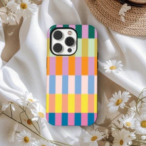 Puede incluir: Funda de teléfono colorida con un patrón de rayas verticales en tonos rosa, naranja, amarillo, azul y verde. La funda tiene un acabado brillante y un módulo de cámara negro. Margaritas y un sombrero de paja en el fondo.