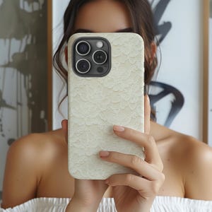 Puede incluir: Una funda de iPhone color crema con un delicado dise&ntilde;o de encaje. La funda tiene un m&oacute;dulo de c&aacute;mara negro con tres lentes y un flash. La funda del tel&eacute;fono se sostiene frente a una persona.