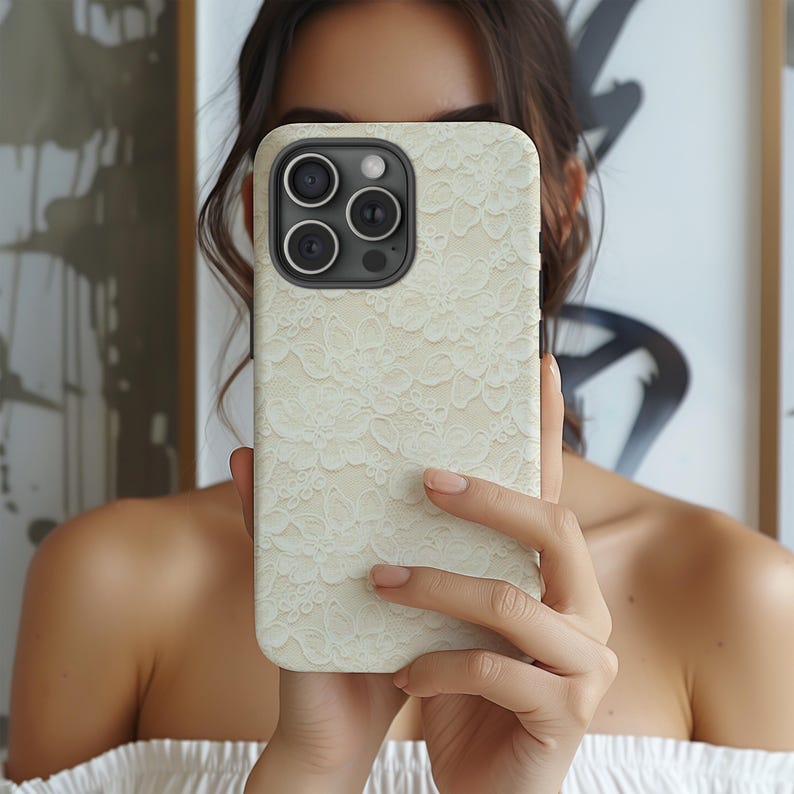 Puede incluir: Una funda de iPhone color crema con un delicado dise&ntilde;o de encaje. La funda tiene un m&oacute;dulo de c&aacute;mara negro con tres lentes y un flash. La funda del tel&eacute;fono se sostiene frente a una persona.