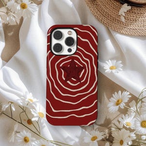 Pode incluir: Capa de telefone vermelha com um design de redemoinho concêntrico branco e uma estrela vermelha escura no centro. A capa tem um módulo de câmera preto. Margaridas brancas estão dispostas ao redor.