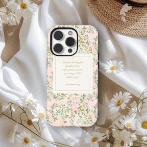 May include: A floral phone case with a pink background and a white and green floral pattern. The case features a rectangular text box with the words "Seek first the kingdom of God and His righteousness, and all these things will be added to you. Matthew 6:33."