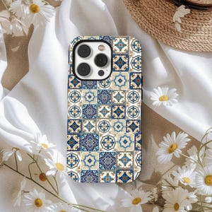 Può includere: Custodia per smartphone con un motivo a piastrelle ripetuto nei toni del blu, bianco e oro. La custodia ha una finitura lucida e un ritaglio per la fotocamera. Il design ricorda le tradizionali piastrelle in ceramica. Margherite e un cappello di paglia sullo sfondo.