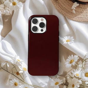 May include: A glossy burgundy phone case. The case features a camera cutout with multiple lenses. The background includes white fabric, daisies, and a straw hat. The case is designed for a smartphone.