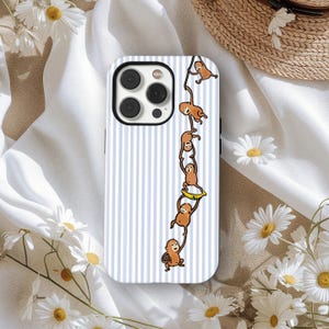 May include: A white and blue striped phone case featuring a cartoon illustration of several brown monkeys hanging from a vine. One monkey holds a yellow banana. The case has a black camera lens.