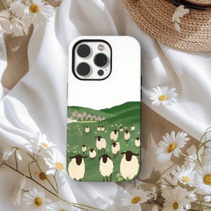 Op de afbeelding: Witte telefoonhoes met een pastorale scène met een kudde schapen die grazen op een groene heuvel onder een witte lucht. De schapen hebben zwarte gezichten en poten. De hoes heeft een zwarte cameralensrand.