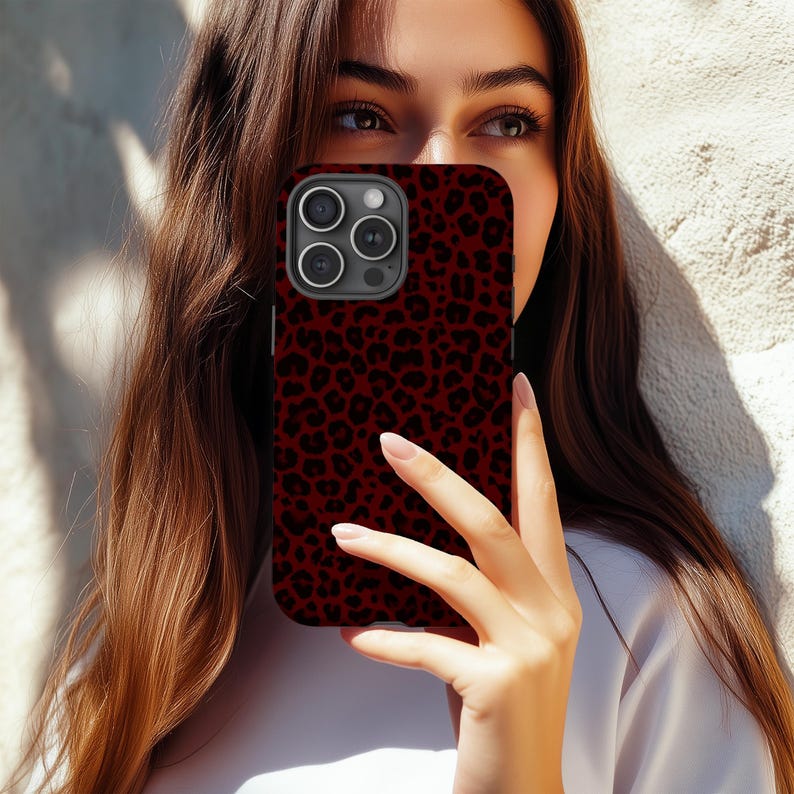 May include: A burgundy phone case with a black leopard print pattern. The case is held in front of a person's face, with the phone's camera lenses visible. The background is a textured, off-white wall.
