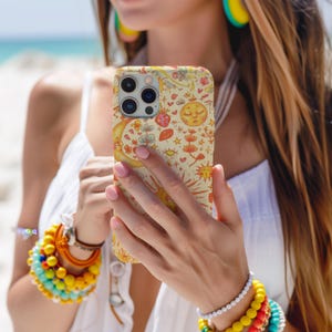 May include: A beige phone case featuring a whimsical celestial design with a crescent moon, sun, stars, and other decorative elements. The case is held in a person's hands, with the beach and ocean visible in the background.