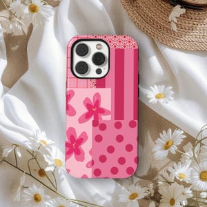 May include: A pink phone case with a patchwork design featuring floral, striped, and polka dot patterns. The case has a camera cutout. Daisies and a straw hat surround the case.