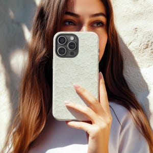 Puede incluir: Funda de tel&eacute;fono blanca con un delicado dise&ntilde;o de encaje. La funda tiene un acabado mate y un recorte para la c&aacute;mara con tres lentes. La funda del tel&eacute;fono se sostiene frente a una persona.
