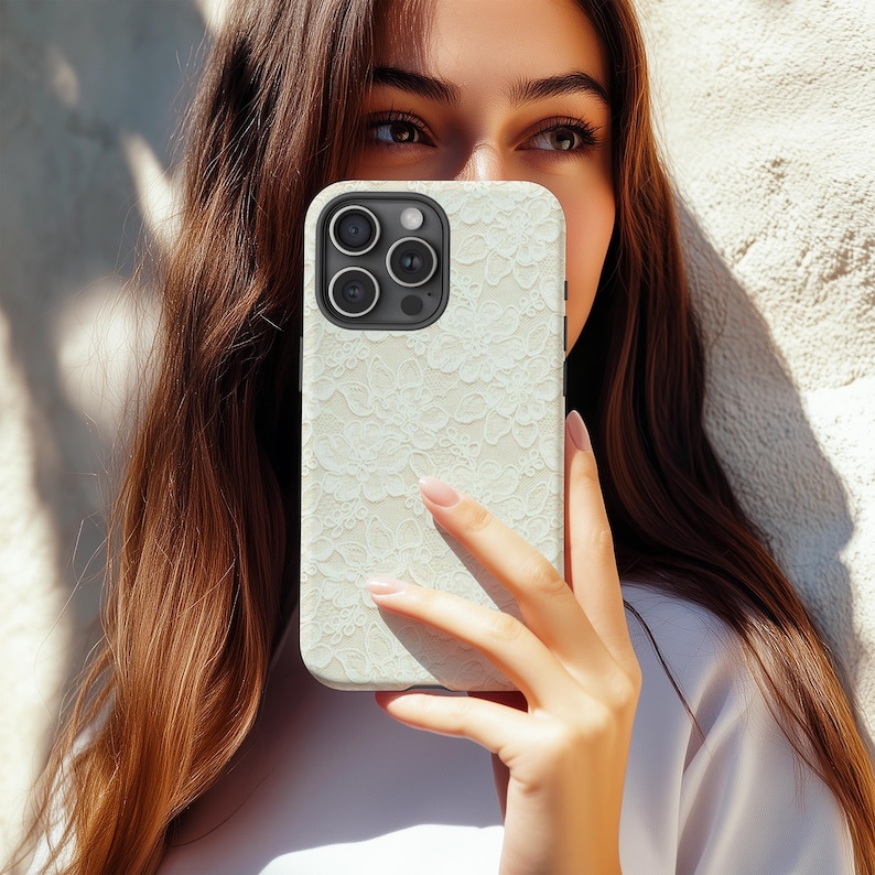 Puede incluir: Funda de tel&eacute;fono blanca con un delicado dise&ntilde;o de encaje. La funda tiene un acabado mate y un recorte para la c&aacute;mara con tres lentes. La funda del tel&eacute;fono se sostiene frente a una persona.