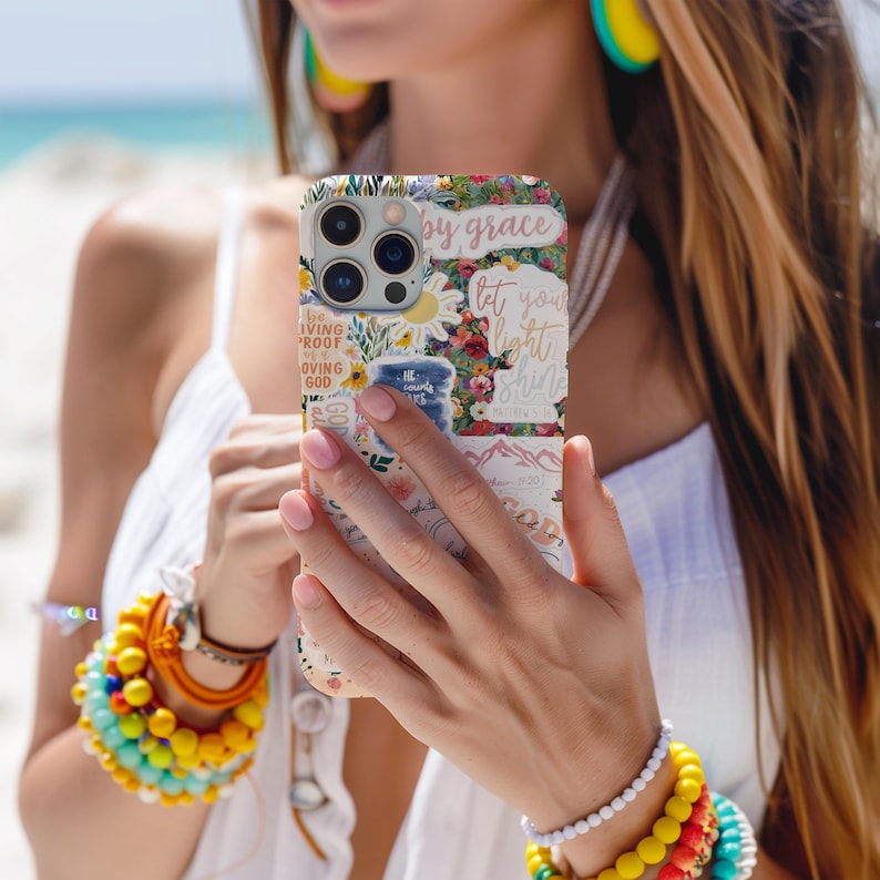 May include: A floral phone case with the words "by grace" and "let your light shine". The case features a colorful design with flowers, mountains, and other decorative elements. The phone case is held by a person.