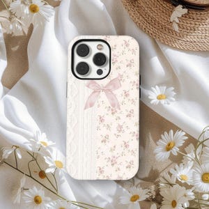 May include: A phone case featuring a cable knit design and a floral pattern. The left side has a cream cable knit, while the right side displays a floral print with pink roses on a cream backdrop. A light pink bow is centered. The case has a black camera module.