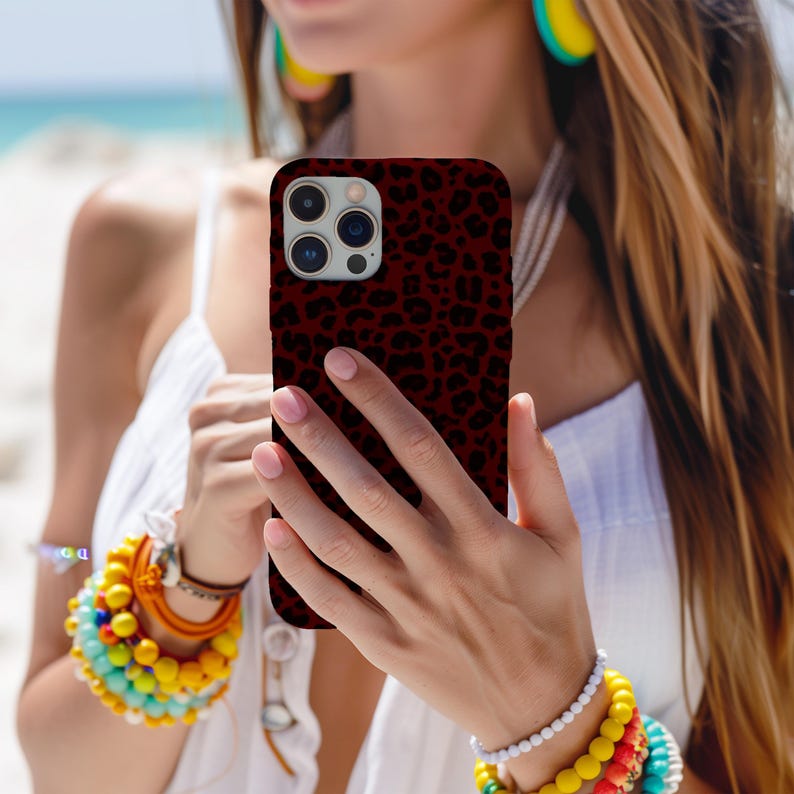 May include: A burgundy phone case with a black leopard print pattern. The case is held in front of a person wearing a white top and colorful bracelets. The background shows a beach scene.