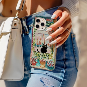 May include: A colorful phone case featuring a black cat illustration. The case has a painted design with a cat, flowers, and a window scene. The case is held by a person wearing blue jeans and a white top.