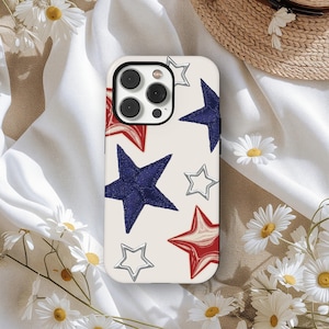 Pode incluir: Uma capa de telefone com fundo creme com um padrão de estrelas vermelhas, azuis e prateadas. A capa tem um recorte para a câmera e é cercada por margaridas brancas e um chapéu de palha.