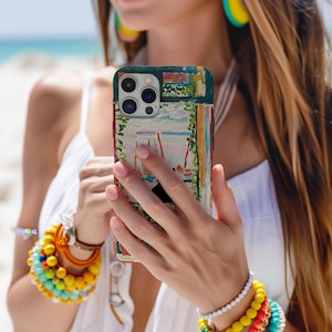 May include: A colorful phone case featuring an impressionistic painting of a seaside scene with sailboats and a cafe. The case has a dark green border and a textured, artistic design. The case is held in front of a beach background.