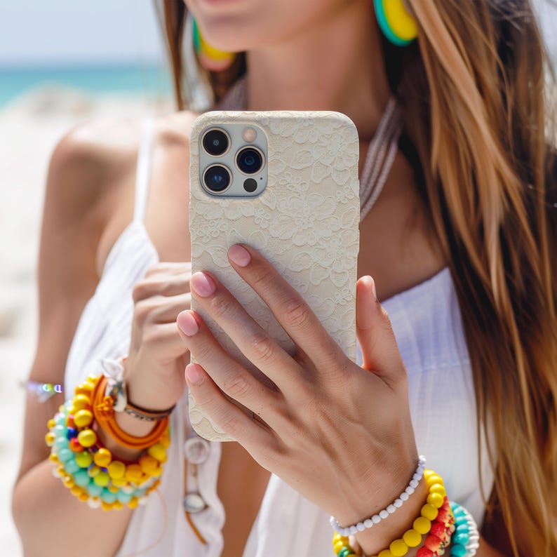 Puede incluir: Funda de tel&eacute;fono de color crema con un delicado dise&ntilde;o de encaje. La funda tiene un recorte para la c&aacute;mara. La persona lleva un vestido blanco y pulseras coloridas. La funda se sostiene frente a la persona.