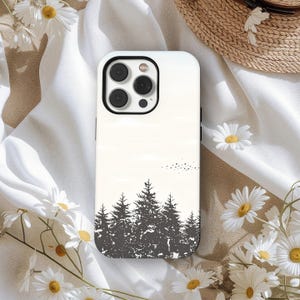 Könnte beinhalten: Weißes Handycase mit schwarzer Waldsilhouette. Das Case hat eine glänzende Oberfläche und einen Kameraausschnitt. Gänseblümchen und ein Strohhut befinden sich im Hintergrund.