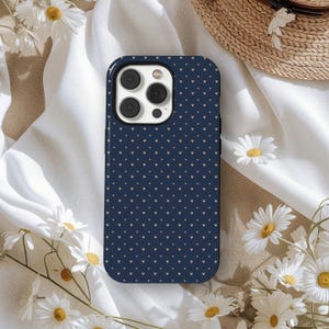 May include: A navy blue phone case with a pattern of small, light-colored hearts. The case has a black border and a camera cutout. The phone case is surrounded by white daisies and a straw hat.