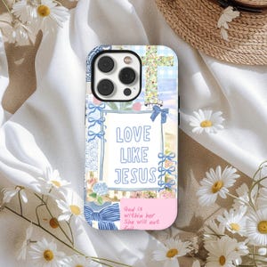 Op de afbeelding: Decoratieve telefoonhoes met de tekst "LOVE LIKE JESUS" in een witte lijst, versierd met blauwe strikken en bloemmotieven. De hoes heeft een kruisontwerp en een roze gedeelte met tekst. Madeliefjes omringen de hoes.
