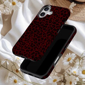 May include: A smartphone case with a burgundy leopard print design. The case is shown in two parts, with the top piece slightly lifted, revealing the phone's camera and screen. The background includes white fabric and daisies.