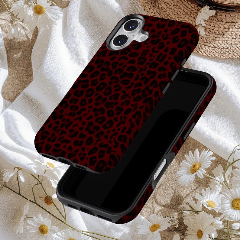 May include: A smartphone case with a burgundy leopard print design. The case is shown in two parts, with the top piece slightly lifted, revealing the phone's camera and screen. The background includes white fabric and daisies.