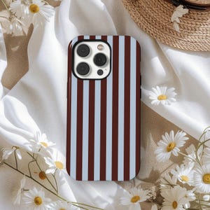 Peut inclure: Coque de smartphone à rayures verticales marron et bleu clair. Le boîtier a un bord noir et une découpe pour l'appareil photo. Le fond comprend du tissu blanc, des marguerites et un chapeau de paille.