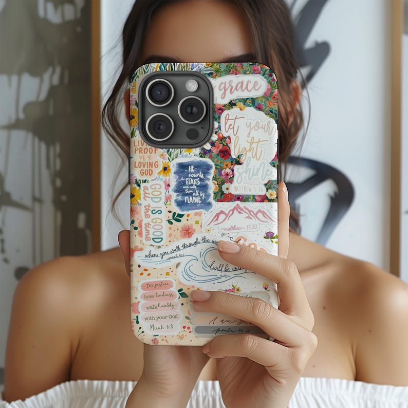 May include: A floral phone case with various inspirational quotes and phrases. The case features a collage of colorful flowers, text that reads "grace", "let your light shine", and other religious phrases. The case is held up in front of a person's face.