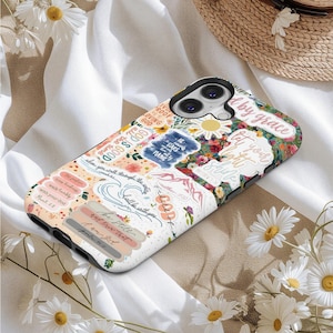 May include: A white and black phone case with a floral and text design. The case features various stickers with inspirational quotes and floral patterns. The text includes phrases like "God is Good" and "Be Still".