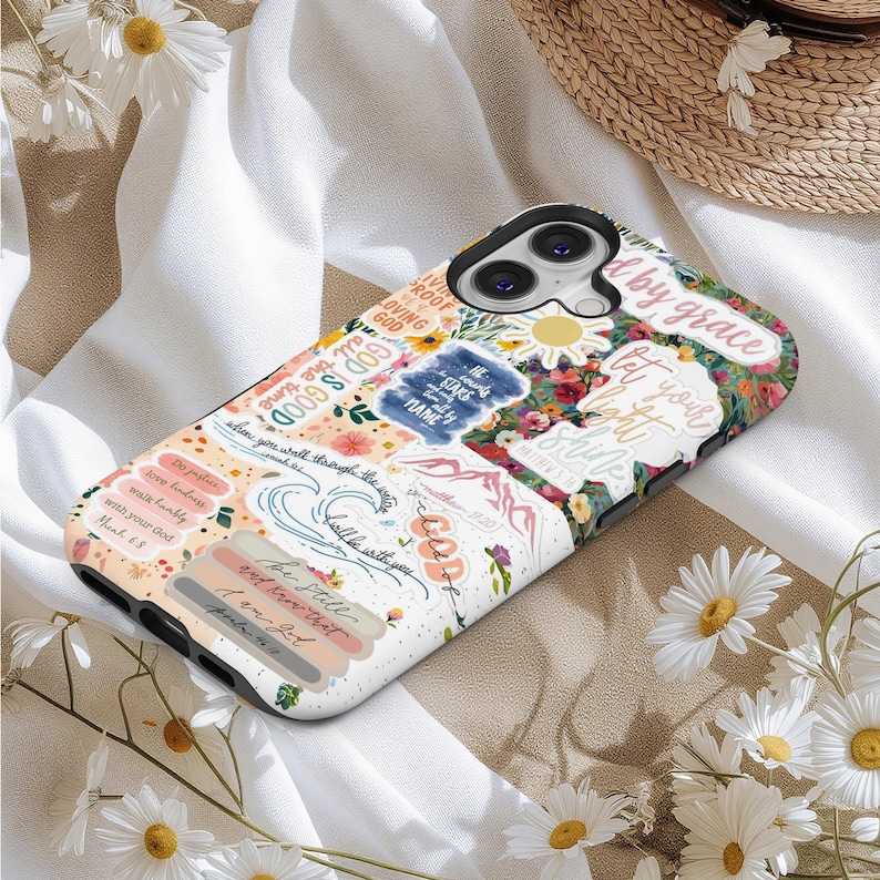 May include: A white and black phone case with a floral and text design. The case features various stickers with inspirational quotes and floral patterns. The text includes phrases like "God is Good" and "Be Still".