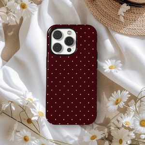 May include: A burgundy phone case with a white polka dot pattern. The case has a black border and a camera cutout. The background includes white fabric, daisies, and a straw hat.