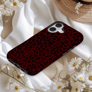 May include: A smartphone case with a dark red leopard print design. The case has a black border and a camera cutout. The case is lying on a white and tan striped surface, surrounded by white flowers and a straw hat.