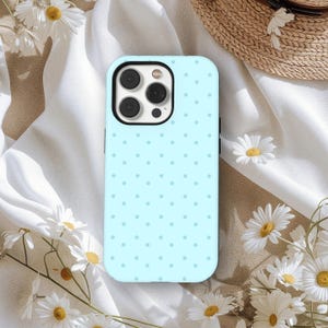 Könnte beinhalten: Hellblaue Handyhülle mit einem Polka-Dot-Muster. Die Hülle hat einen schwarzen Kameraobjektiv-Ausschnitt und ist von weißen Gänseblümchen und einem Strohhut umgeben.
