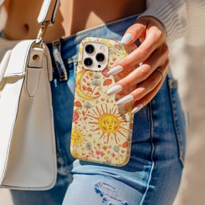 May include: A phone case with a whimsical design featuring suns, moons, stars, and flowers in shades of yellow, orange, and red. The case is held by a person wearing blue jeans and a white sweater. A white purse is visible in the background.