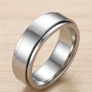 Può includere: Un anello argentato lucido con una superficie liscia e lucida. L'anello presenta un design semplice e moderno con una fascia centrale che sembra ruotare liberamente. Realizzato in metallo riflettente, probabilmente acciaio inossidabile.