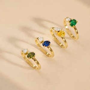 Könnte beinhalten: Vier Goldringe, die in einer diagonalen Linie angeordnet sind. Jeder Ring verfügt über einen ovalen Edelstein in Krappenfassung und einen kleineren, birnenförmigen, klaren Stein. Die Edelsteine sind olivgrün, tiefblau, leuchtend gelb und smaragdgrün.