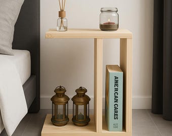 May include: A light-colored wooden side table with a rectangular design. It features a shelf with two small lanterns, a book titled "AMERICAN CARES", and a glass jar with a candle. A reed diffuser sits on the top shelf. The table is next to a bed.