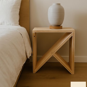 Może przedstawiać: Jasny drewniany stolik nocny o geometrycznym kształcie, z jasnoszarą wazą na górze. Wazon stoi na okrągłej drewnianej podstawie. Stolik nocny stoi obok łóżka z białą pościelą.