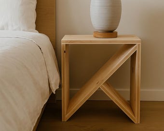 May include: A light wood nightstand with a geometric design, featuring a light gray vase on top. The vase sits on a round wooden base. The nightstand is next to a bed with white bedding.