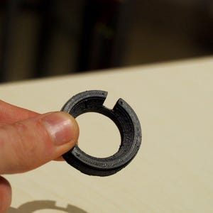 May include: A black, circular 3D-printed object with a textured surface and a slot cut into the side. The object is held in a person's hand, with a neutral background.