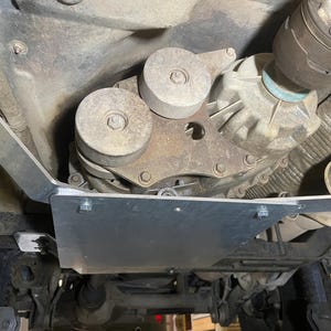 May include: Underside view of a vehicle's drivetrain components. The image shows a transfer case with two circular pulleys, a metal skid plate, and various mechanical parts. The components are bolted together, and the overall color scheme is metallic and gray.