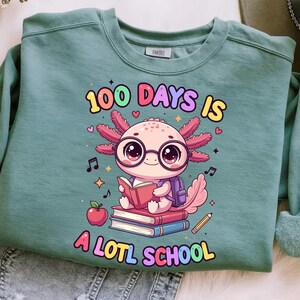 May include: A teal sweatshirt with a colorful graphic that reads "100 DAYS IS A LOTL SCHOOL." The graphic features a cartoon axolotl wearing glasses and a backpack, reading a book on top of a stack of books, with an apple and pencil.