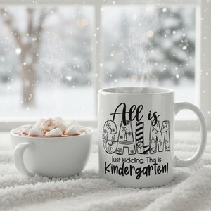 May include: A white ceramic mug with the text "All is Calm, Just kidding. This is Kindergarten!" next to a white bowl of hot chocolate with marshmallows. The mug has a handle and is on a white, fluffy surface.