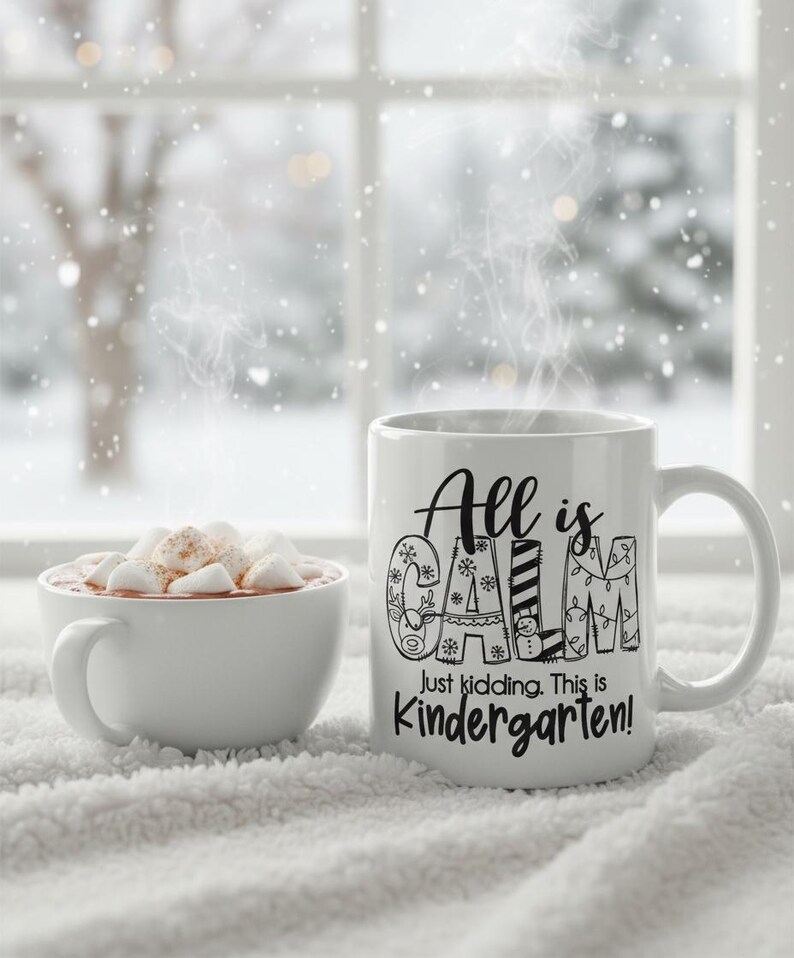 May include: A white ceramic mug with the text "All is Calm, Just kidding. This is Kindergarten!" next to a white bowl of hot chocolate with marshmallows. The mug has a handle and is on a white, fluffy surface.