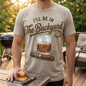 May include: A beige short-sleeve t-shirt featuring the brown text I'LL BE IN The Backyard above a graphic illustration of a crystal glass filled with amber liquid and ice cubes, positioned directly above a brown cigar with a silver band.