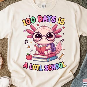 May include: Cream-colored t-shirt featuring a cartoon axolotl wearing glasses and reading a book. The text "100 DAYS IS A LOTL SCHOOL" is printed above and below the axolotl in colorful letters. An apple, pencil, and musical notes are also included.