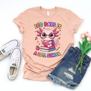 May include: A peach-colored t-shirt with the text "100 DAYS IS A LOTL SCHOOL" and a cartoon axolotl wearing glasses and reading a book. The image includes white sneakers and denim shorts.